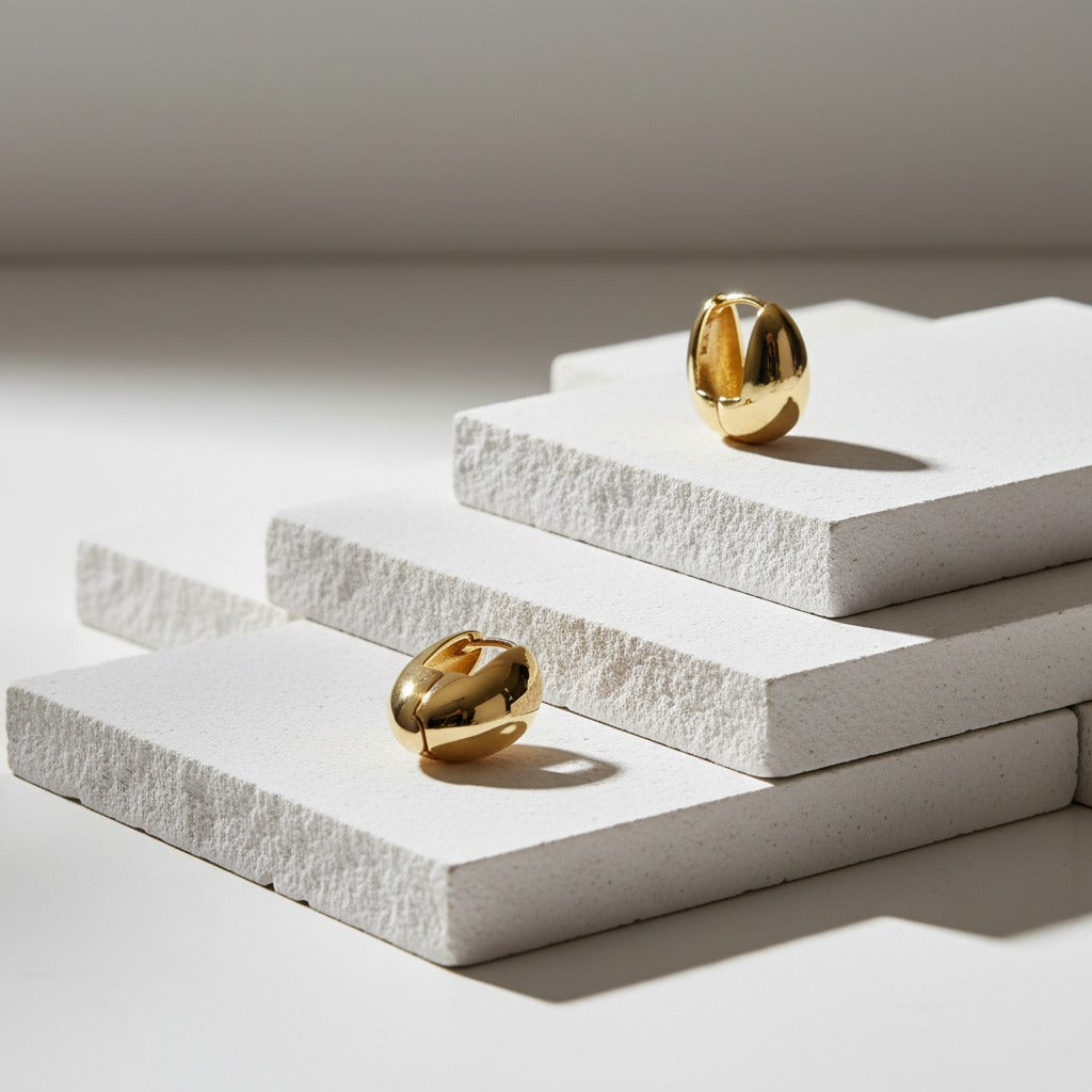 Two gold earrings on white stone plinths with a neutral background
