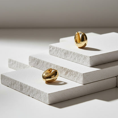 Two gold earrings on white stone plinths with a neutral background