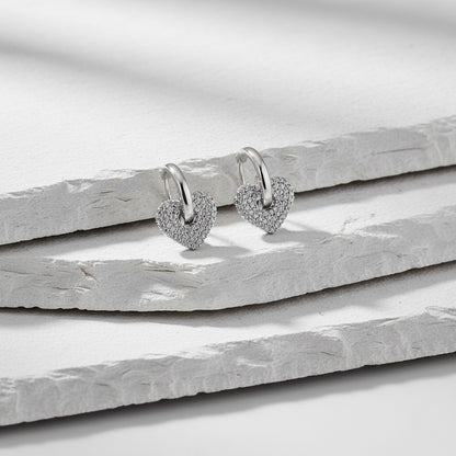 Heart-shaped silver earrings with small diamonds on a light wooden surface