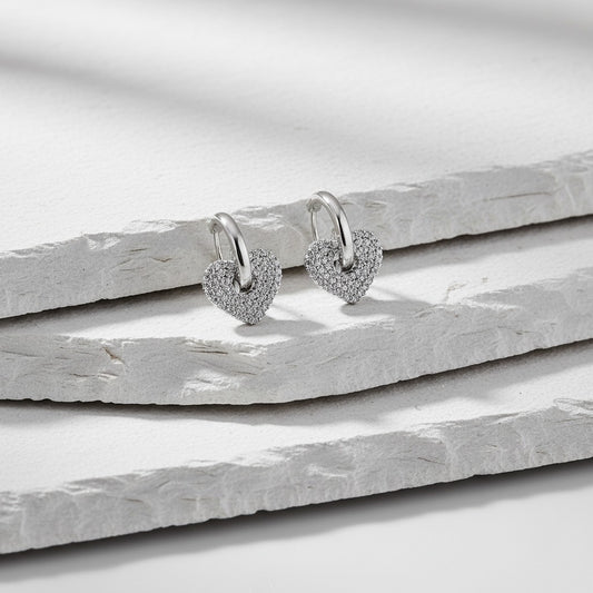 Heart-shaped silver earrings with small diamonds on a light wooden surface
